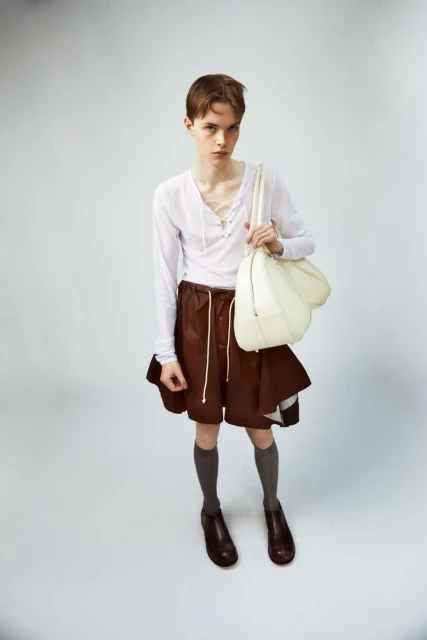 A person poses in a white long-sleeved shirt and brown shorts, carrying a white bag against a pale grey background in a studio setting.