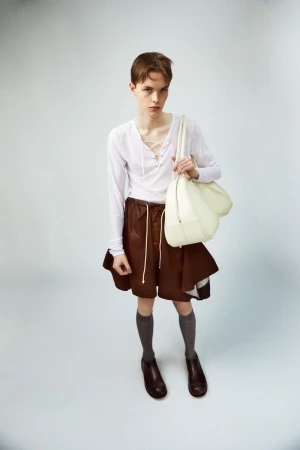 A person poses in a white long-sleeved shirt and brown shorts, carrying a white bag against a pale grey background in a studio setting.