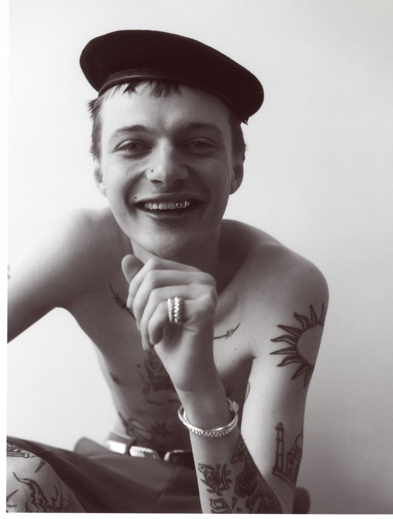 Black and white portrait of a young adult man wearing a beret with tattoos, jewelry, and a tooth grill, smiling in a studio setting.