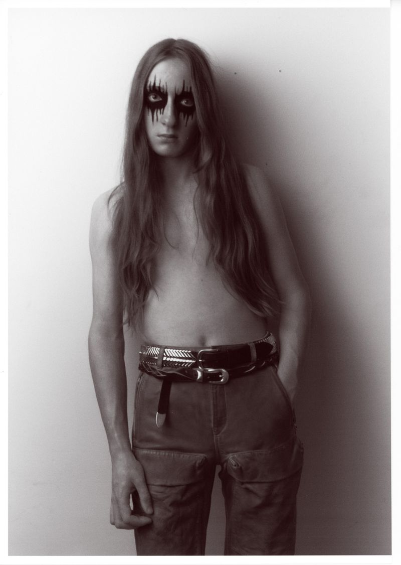 Portrait of a topless male with long brown hair and black eye makeup, wearing pants and two belts, standing indoors against a white wall.