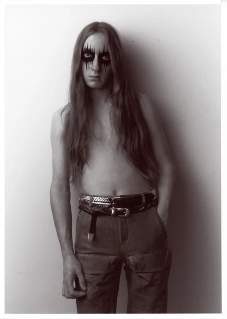 Portrait of a topless male with long brown hair and black eye makeup, wearing pants and two belts, standing indoors against a white wall.
