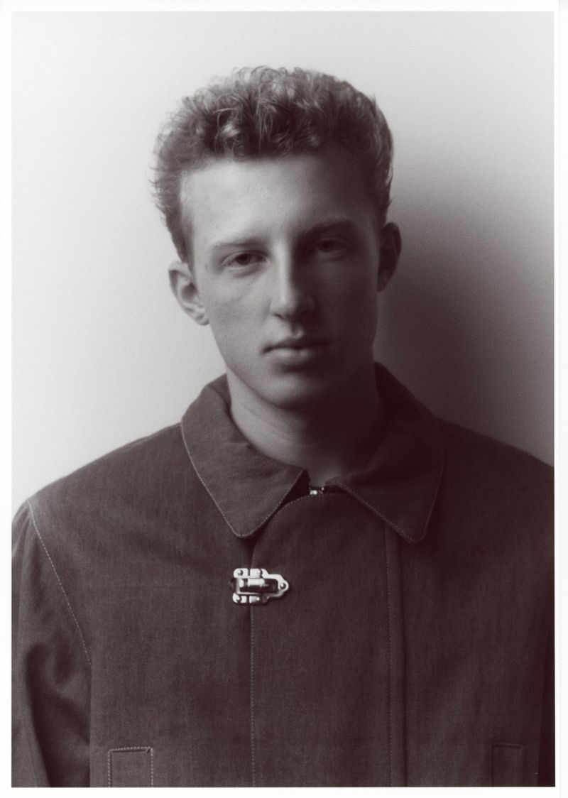Monochrome portrait of a young adult male with short curly hair wearing a collared jacket in a studio setting against a neutral background.