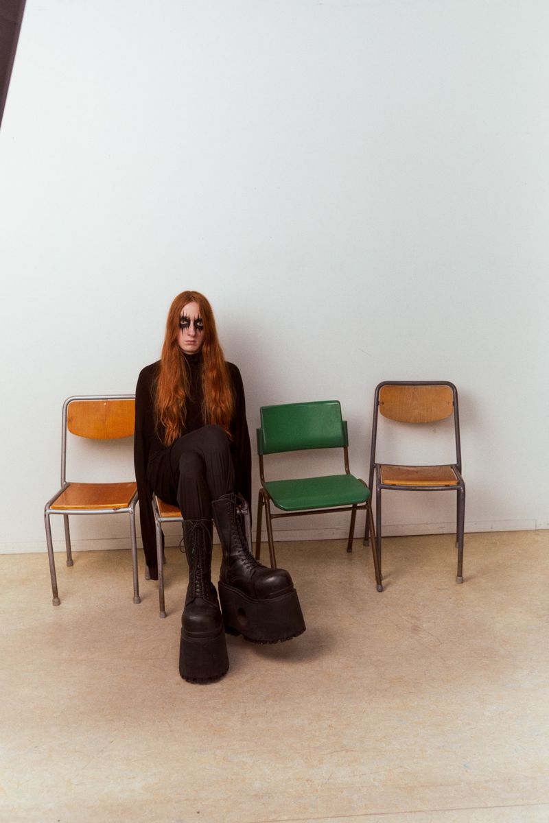 Woman with long red hair wears black sunglasses and a black outfit with platform boots sitting on an orange chair in a photo studio.