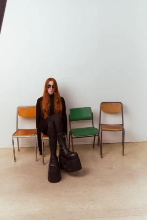 Woman with long red hair wears black sunglasses and a black outfit with platform boots sitting on an orange chair in a photo studio.