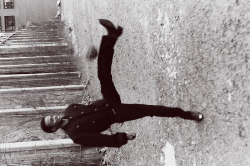 Monochrome shot of adult performing acrobatic kick in mid-air against weathered wall with stairs visible in the upper left.