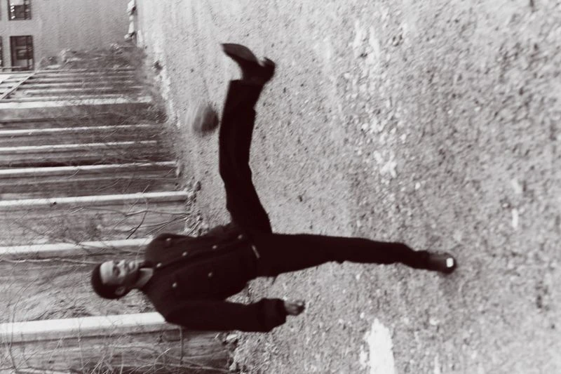 Monochrome shot of adult performing acrobatic kick in mid-air against weathered wall with stairs visible in the upper left.
