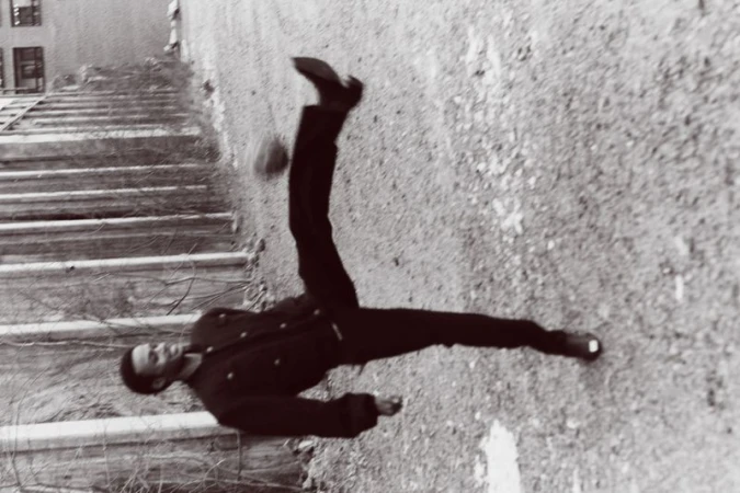 Monochrome shot of adult performing acrobatic kick in mid-air against weathered wall with stairs visible in the upper left.