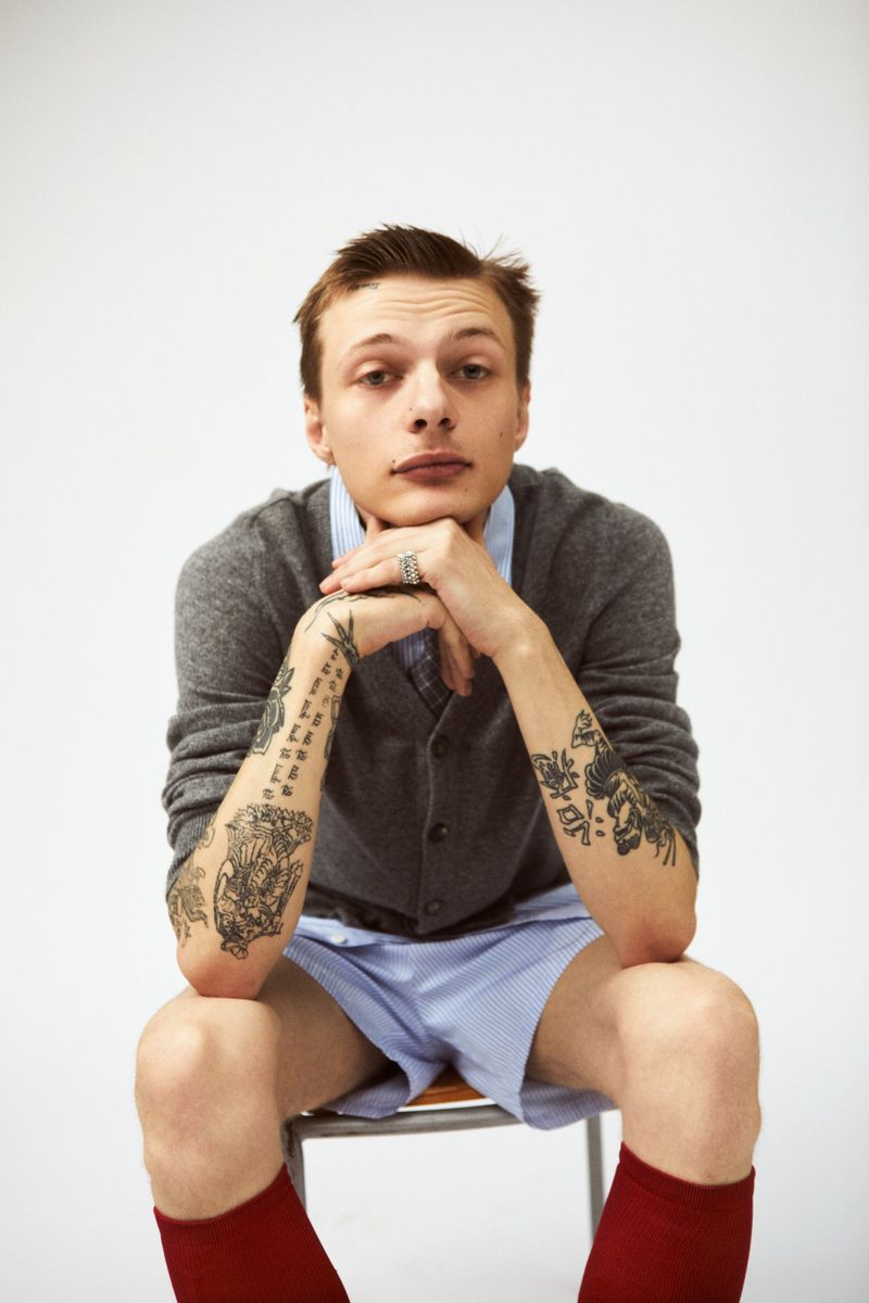 Young man with tattooed arms seated on a chair wearing a grey cardigan and blue shorts against a white backdrop