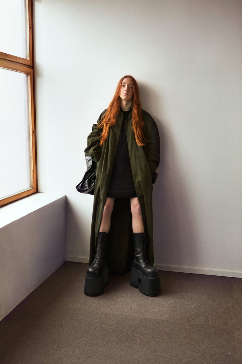 A long-haired person in platform boots and a coat stands in a corner with white walls and a window.