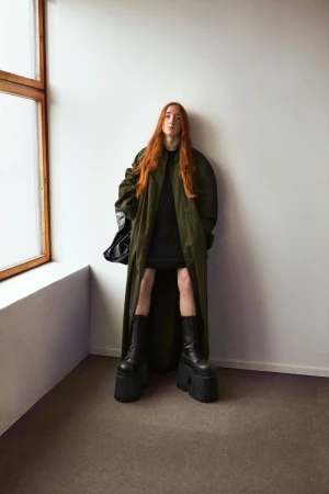 A long-haired person in platform boots and a coat stands in a corner with white walls and a window.