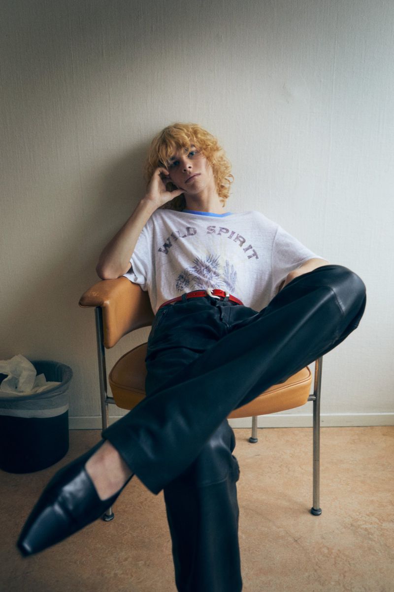 A young adult with curly blonde hair sits in a chair wearing a graphic t-shirt, dark pants, and black loafers in an indoor setting.
