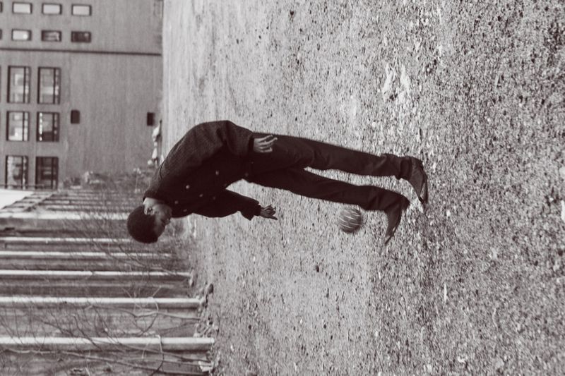 Monochrome photograph of a young man playing with a ball in an outdoor urban setting, showing buildings and a sidewalk.