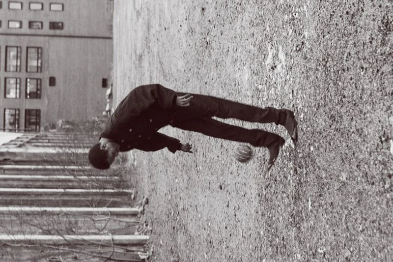 Monochrome photograph of a young man playing with a ball in an outdoor urban setting, showing buildings and a sidewalk.