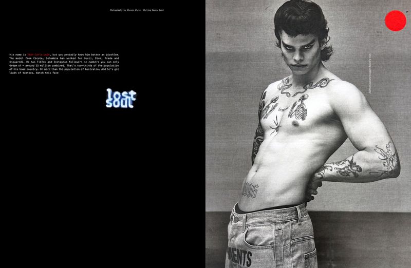 Editorial shot of a shirtless adult male model with tattoos, wearing light-wash jeans, posing against a grey background with a red circle.