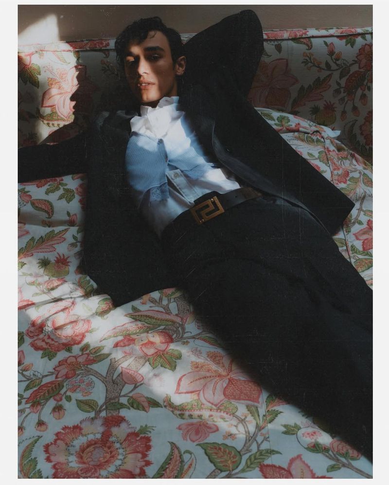 Adult male model in black suit and white ruffled shirt reclining on floral sofa with sunlight casting shadows in indoor setting.