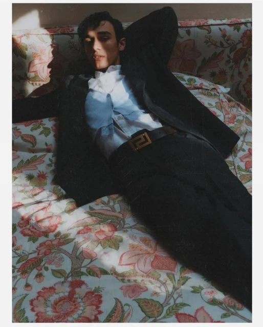 Adult male model in black suit and white ruffled shirt reclining on floral sofa with sunlight casting shadows in indoor setting.