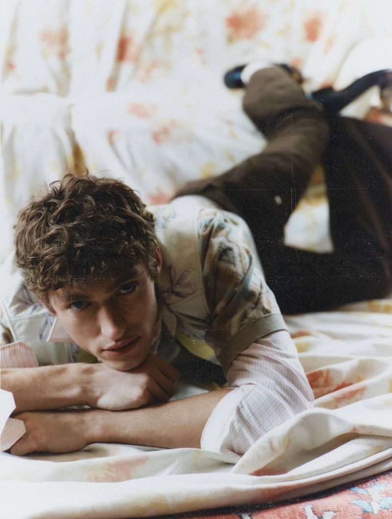 Young adult male laying on patterned bedding, wearing a floral print shirt. Indoor setting, eye-level angle, soft lighting.