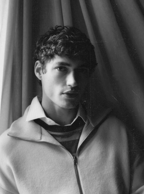 Monochromatic portrait of a young adult male with curly hair, wearing a jacket and collared shirt, posing in front of draped curtains.