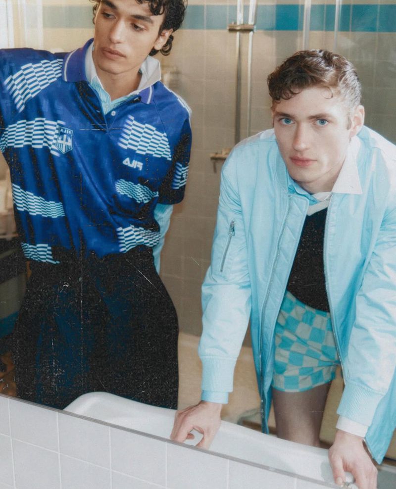 Two young adult men in a bathroom with cool-toned fashion. One wears a blue polo, the other a light blue bomber jacket. Indoor setting.