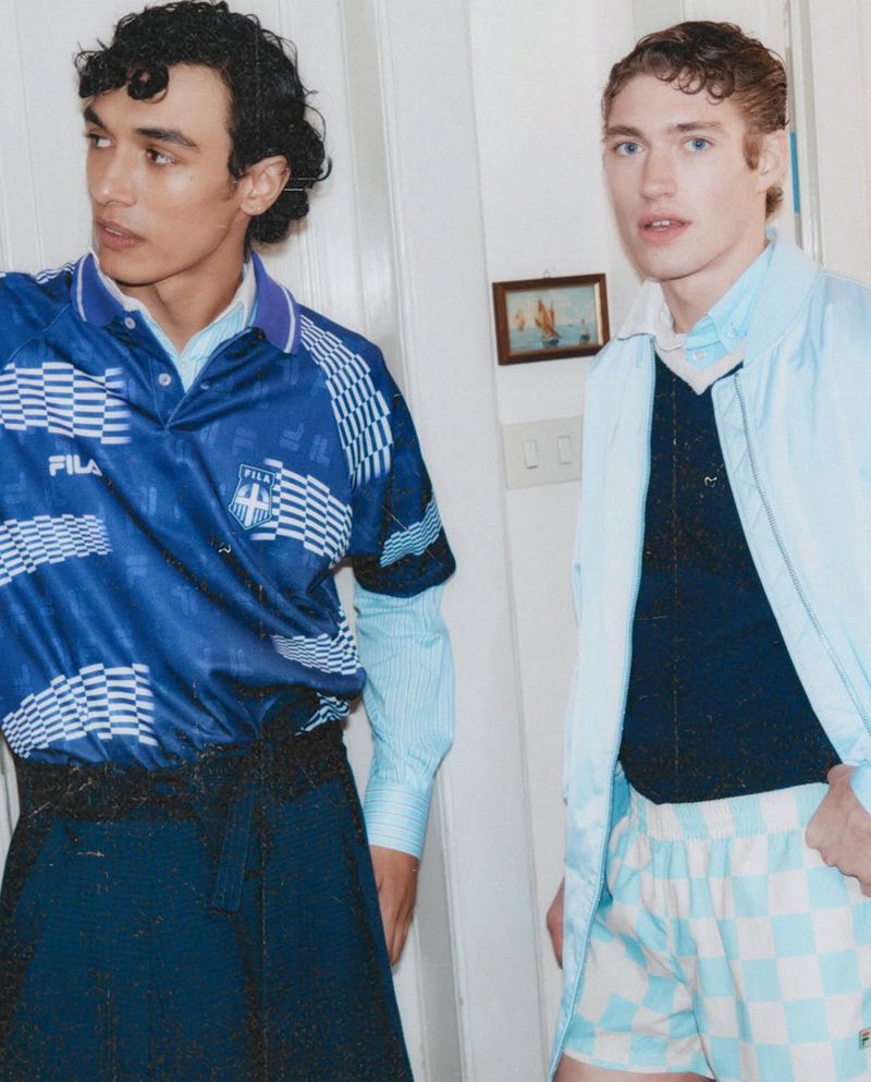 Two men pose indoors wearing a blue soccer jersey, navy sweater, blue shorts, and a light blue jacket. The backdrop is a white wall.