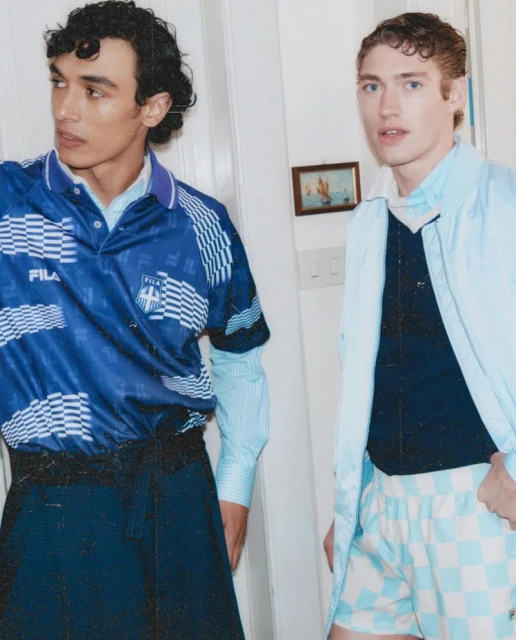 Two men pose indoors wearing a blue soccer jersey, navy sweater, blue shorts, and a light blue jacket. The backdrop is a white wall.