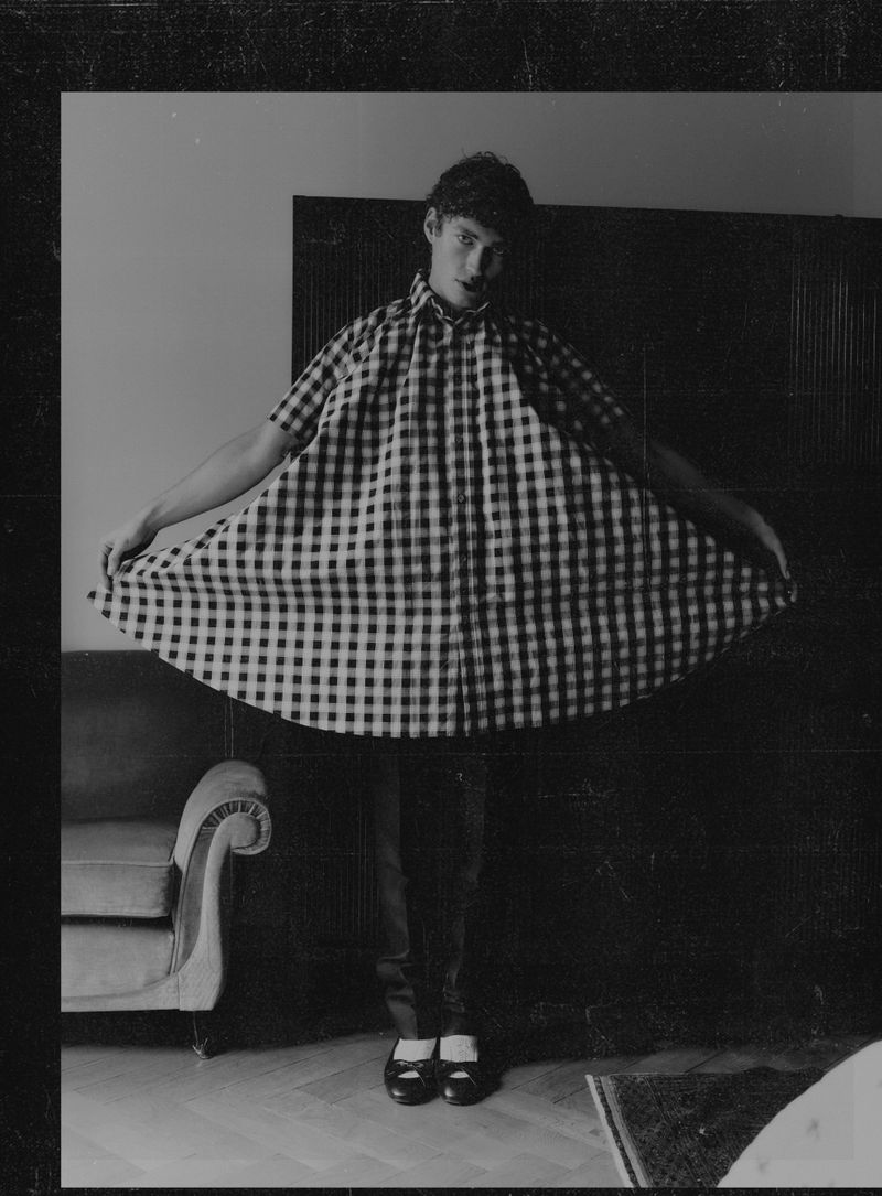 Black and white portrait of a young adult male wearing a checkered oversized shirt and black pants indoors, styled in a fashion-forward manner.
