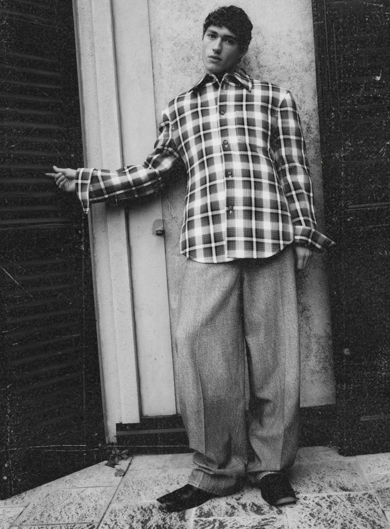 Monochrome portrait of a male model wearing a patterned plaid shirt and wide trousers standing near dark shutters.