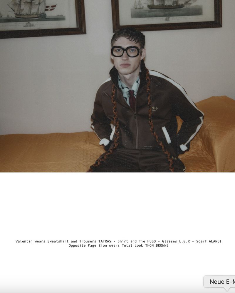Young adult in a brown tracksuit and glasses sits on an orange couch in front of framed artwork featuring vintage ships.
