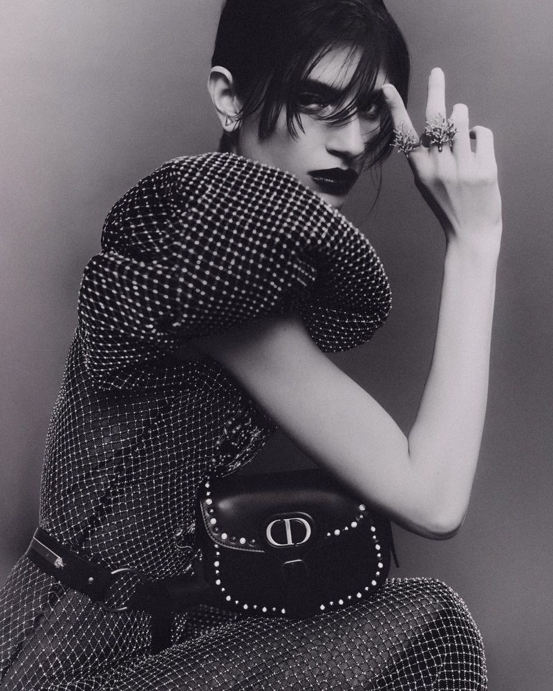 Black-and-white studio portrait of a young adult with dark hair, wearing a patterned top and black bag, posing expressively with jewelry.