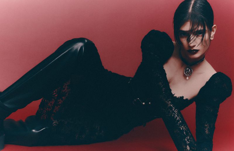 A woman with dark hair and makeup lies on a red surface wearing black lace top and leggings, captured in a dramatic pose and studio lighting.