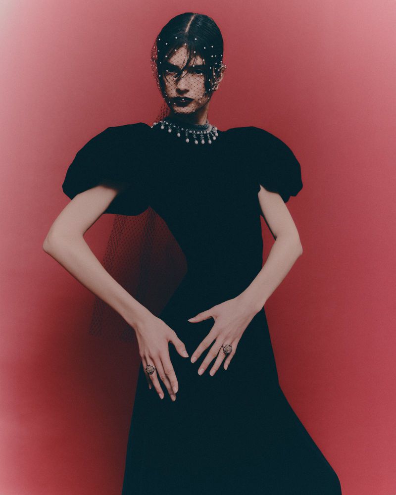 Woman in black gown with pearl necklace and veil poses against pink backdrop, hands near waist, showcasing rings. Elegant fashion photography.