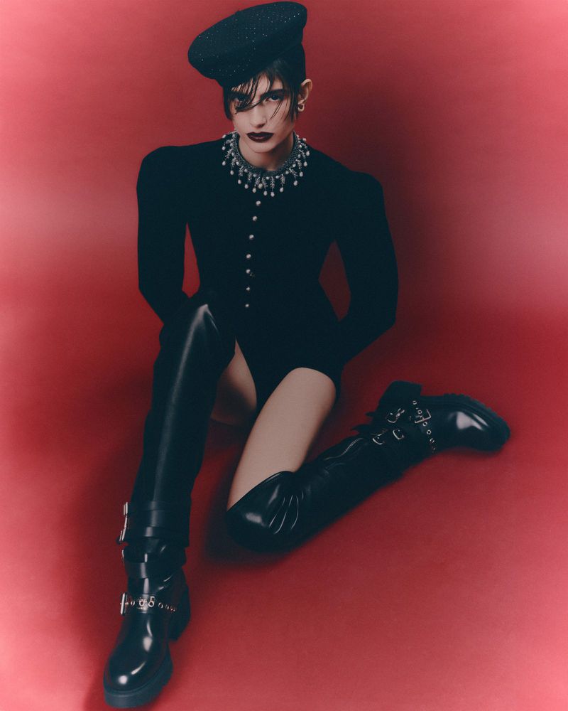 Fashion photo of a female model with dark hair wearing a black bodysuit, beret, necklace, and thigh-high boots sitting in a red studio.