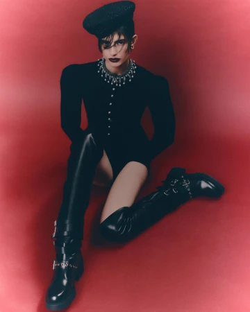 Fashion photo of a female model with dark hair wearing a black bodysuit, beret, necklace, and thigh-high boots sitting in a red studio.