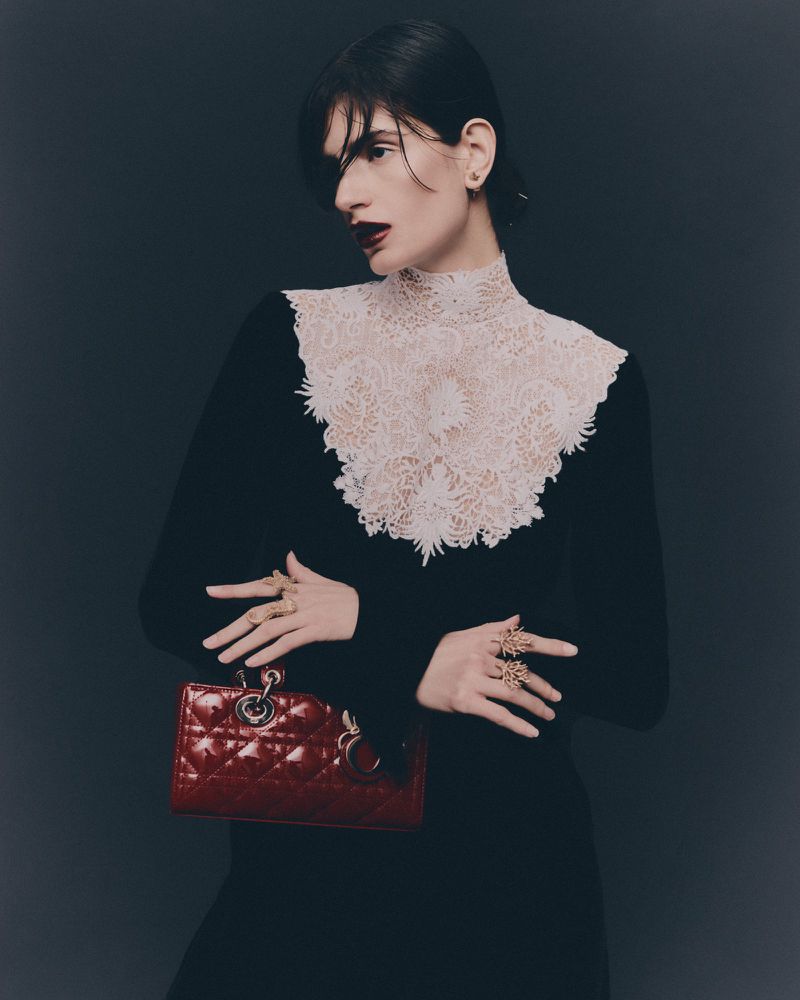 Elegant portrait of a woman in a black dress with lace collar, holding a quilted red purse against a dark background.