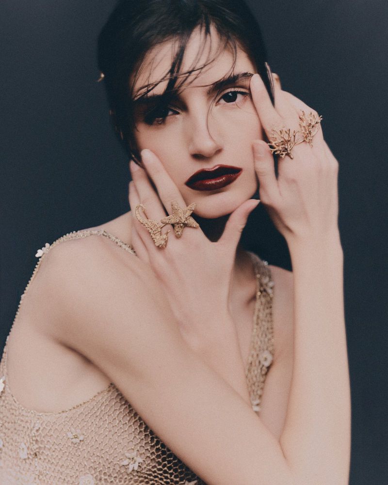 A woman with dark hair and pale skin poses with her hands near her face, wearing rings and a beaded tank top against a dark background.