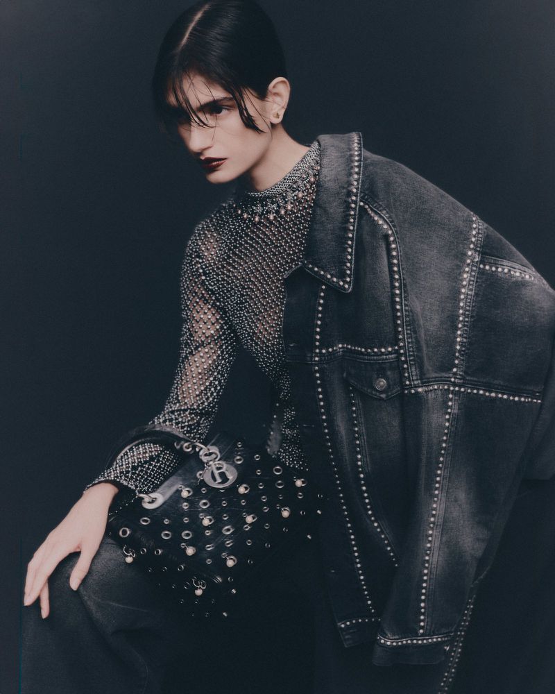 Fashion shot of woman in embellished top and denim jacket holding studded purse in dark studio, eyes downcast, editorial lighting.