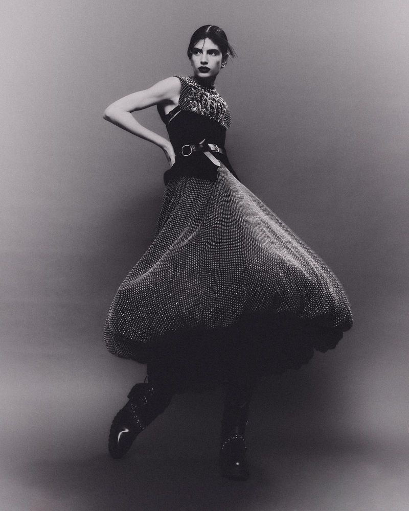 A fashion image in black and white features a female model posing with a voluminous dress, bodice, and boots against a grey backdrop.