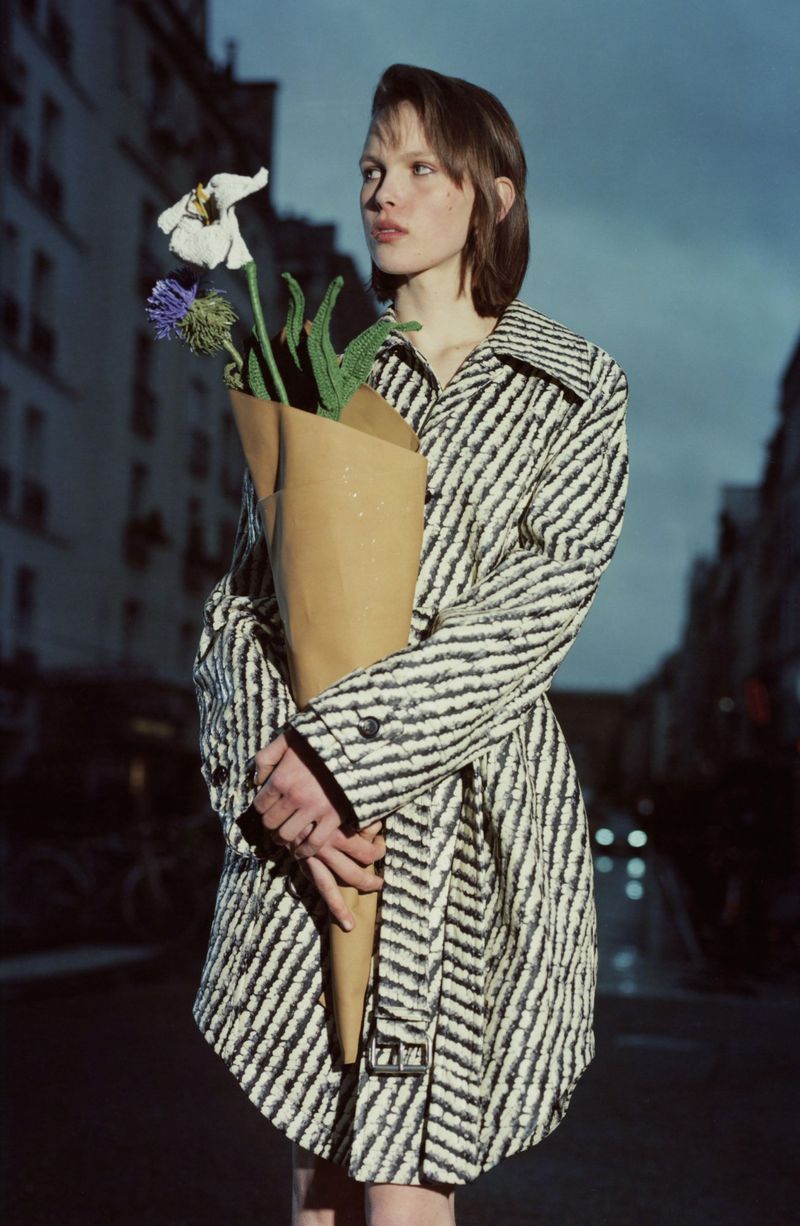 Woman in patterned coat holds a paper-wrapped bouquet of flowers on an urban street at dusk in a fashion editorial image.