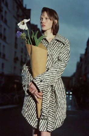 Woman in patterned coat holds a paper-wrapped bouquet of flowers on an urban street at dusk in a fashion editorial image.