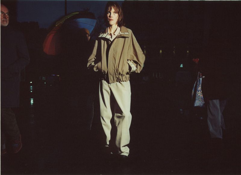 Model in a tan jacket and trousers standing on a dark nighttime street with passing pedestrians and umbrellas