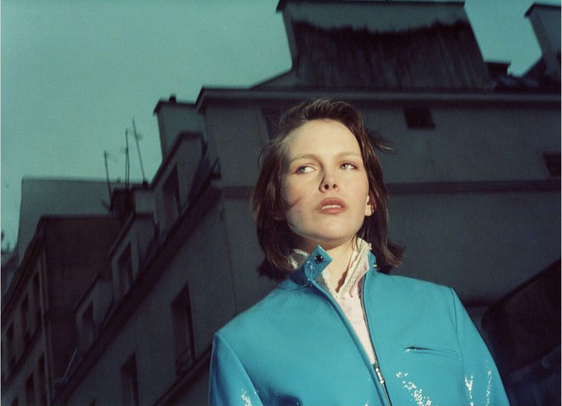 Low-angle view of a young woman in a turquoise jacket against a background of blurred, weathered buildings and a muted, atmospheric sky.