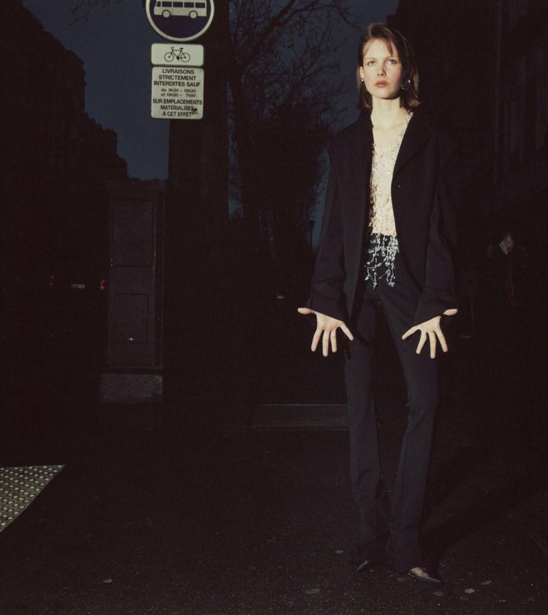 Young adult woman in a dark-blue blazer and pants poses outdoors in the evening, next to a city bus-route sign and a tree.