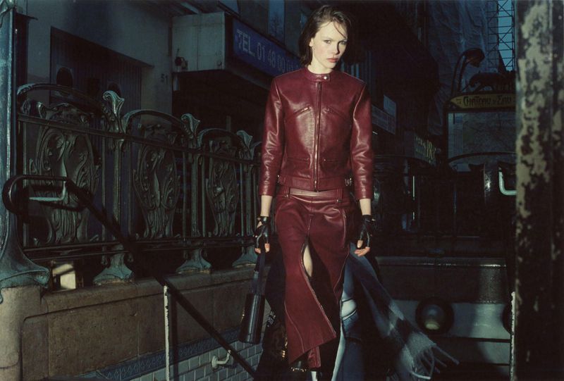 Model Thea Almqvist wearing a dark red leather jacket and matching skirt standing near an outdoor metal railing at dusk.