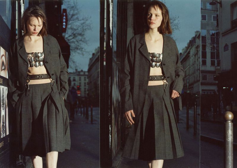 A woman poses in a grey skirt suit and metal bodice on a city street during dusk, with buildings and a 'TABAC' sign in the background.