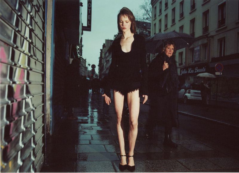 Model Thea Almqvist walking down a wet Parisian street in black attire with a passerby holding an umbrella