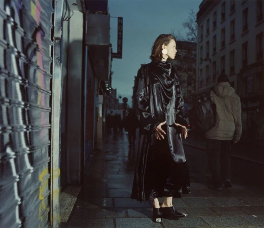 Woman in black coat stands on a wet city street at night. Building and storefronts line the background in an urban scene.