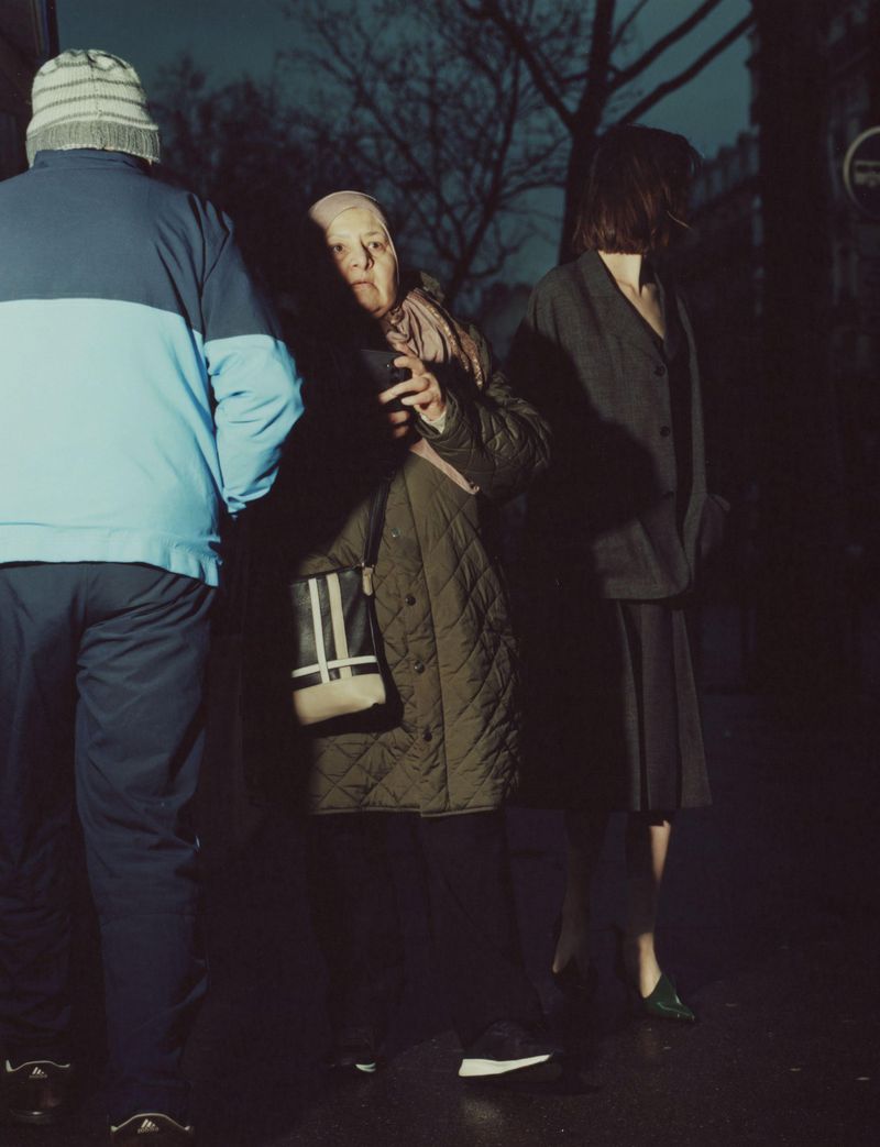 Three people stand on a dark city street at night. An adult male, an elderly female and an adult female stand near each other.