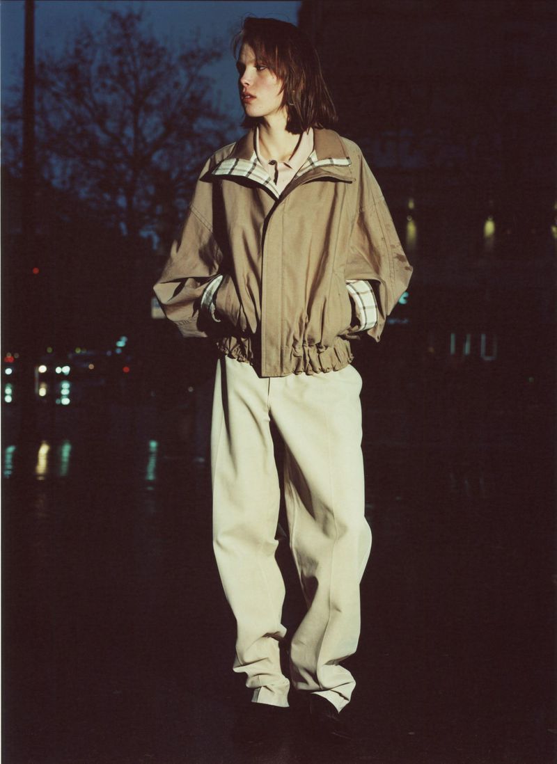 A young adult poses at night in beige jacket and pants, illuminated by city lights reflecting on wet pavement; fashion photography.