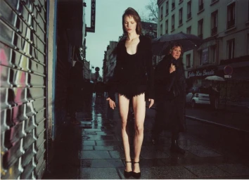 Model Thea Almqvist walking down a wet Parisian street in black attire with a passerby holding an umbrella
