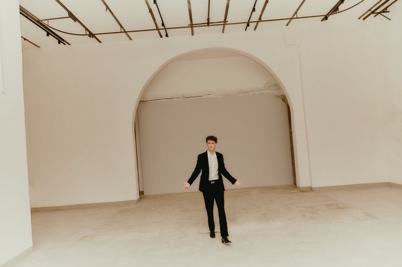Man in a black suit stands in a large, empty room with white walls and arched doorway, looking directly at the camera with arms outstretched.
